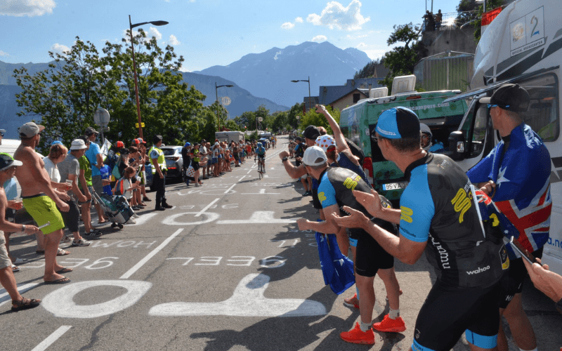 Cyclist racing on road lined with cheering spectators and Mummu Cycling staff.