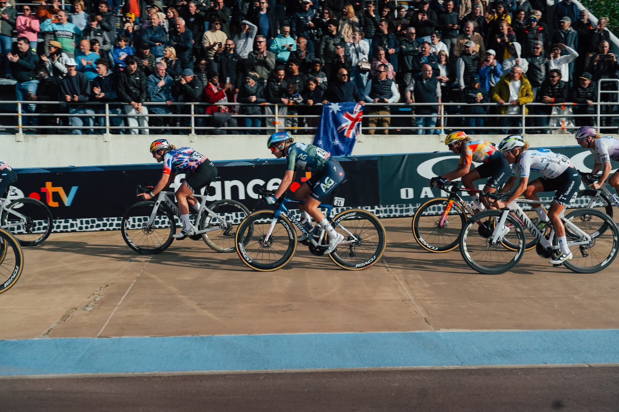 Female cyclists from AG Insurance – Soudal Team and EF Education-EasyPost race intensely on a velodrome track, with spectators and an Australian flag in the background.
