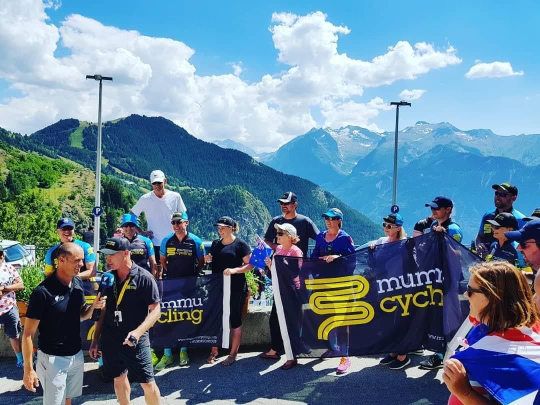 Mummu Cycling group with banner against mountain backdrop.