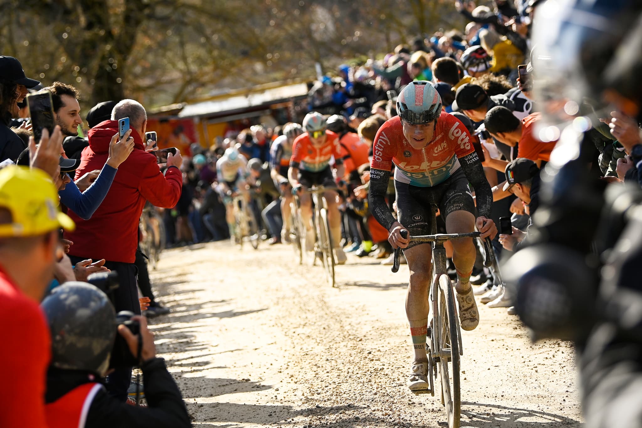 A professional cyclist attacks on the iconic white gravel roads of Strade Bianche, with spectators lining both sides of the dusty strada bianca and a chasing group of riders visible behind.