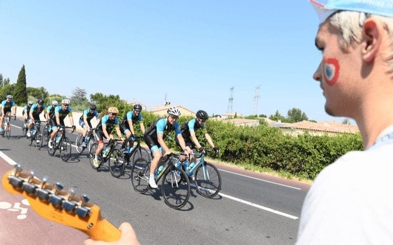 Cyclists ride past a fan with face paint and a guitar.