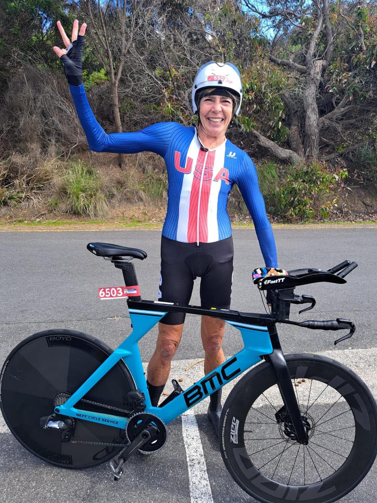 An older woman in a USA national cycling kit smiles and waves next to her BMC time trial bike with race number 6503.