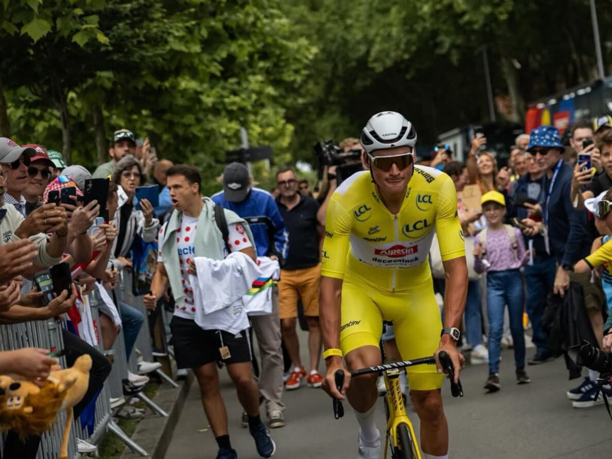 Mathieu van der Poel rides through a crowd of cheering fans while wearing the Tour de France yellow jersey and full yellow kit, surrounded by excited spectators capturing the moment on their phones.